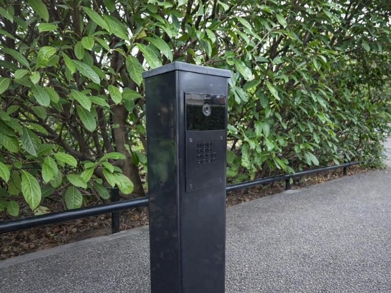 Zwarte zuil met ingebouwde video-intercom en toetsenpaneel langs een pad, omringd door groene struiken.