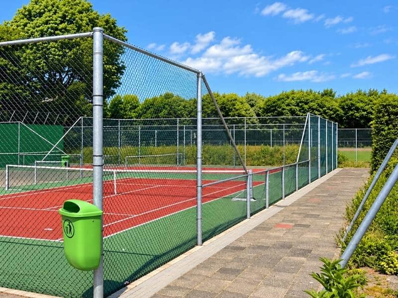 Afrastering tennisbaan met hoog gaashekwerk rondom een rode tennisbaan en groene speelvelden.