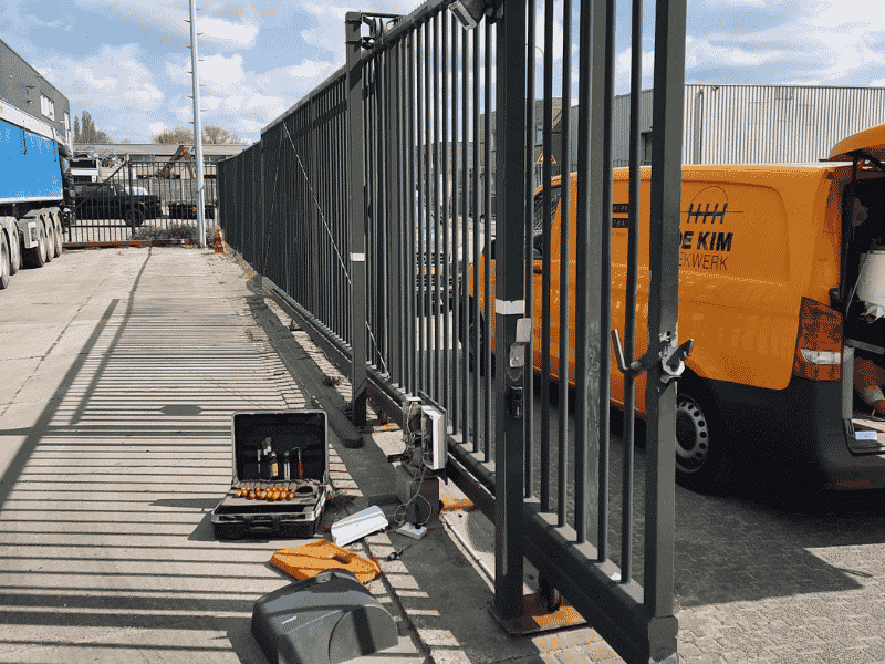 Onderhoud poorten aan een grote elektrische schuifpoort op een bedrijventerrein, met gedemonteerde poortmotor en gereedschap.