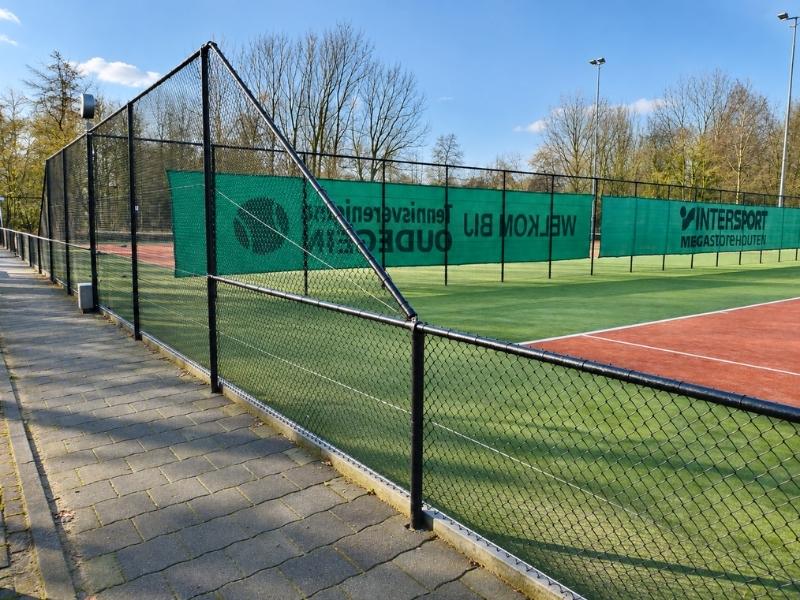 Hekwerk tennisbaan met zwart gaashekwerk en hoge ballenvanger rondom een kunstgras tennisveld